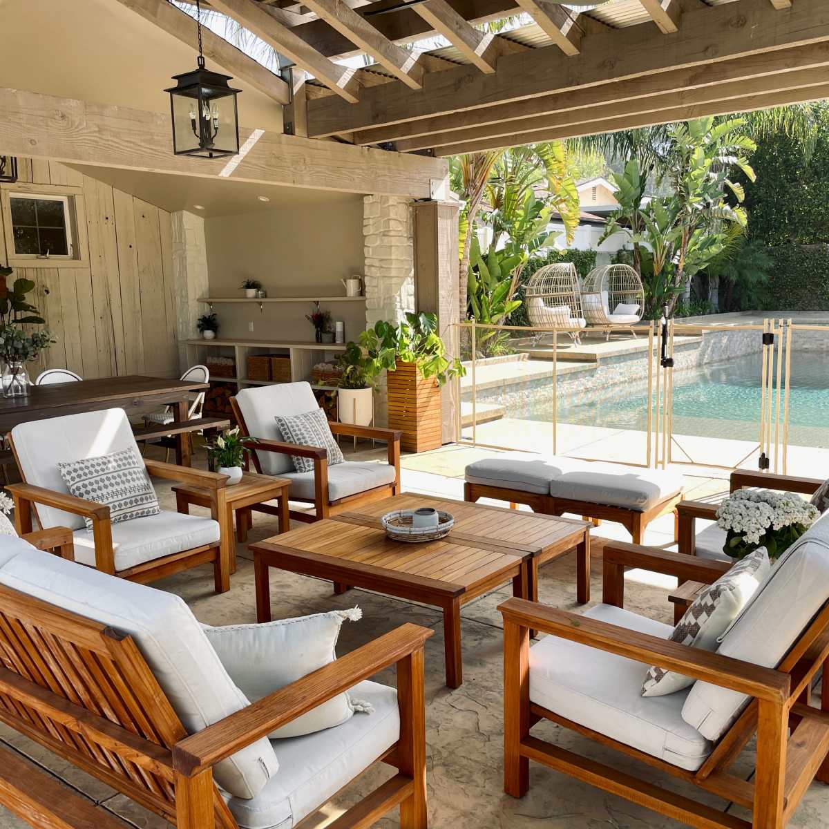 A spacious outdoor seating area features wooden furniture arranged around a central table, overlooking a pool surrounded by greenery.