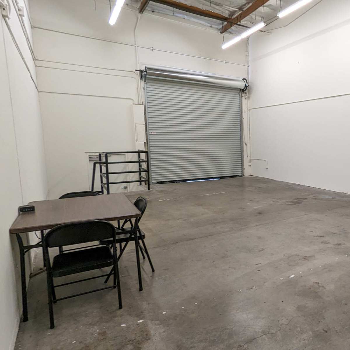 A sparsely furnished room with a concrete floor, a table and chairs, a closed garage door, and several standing light fixture stands.