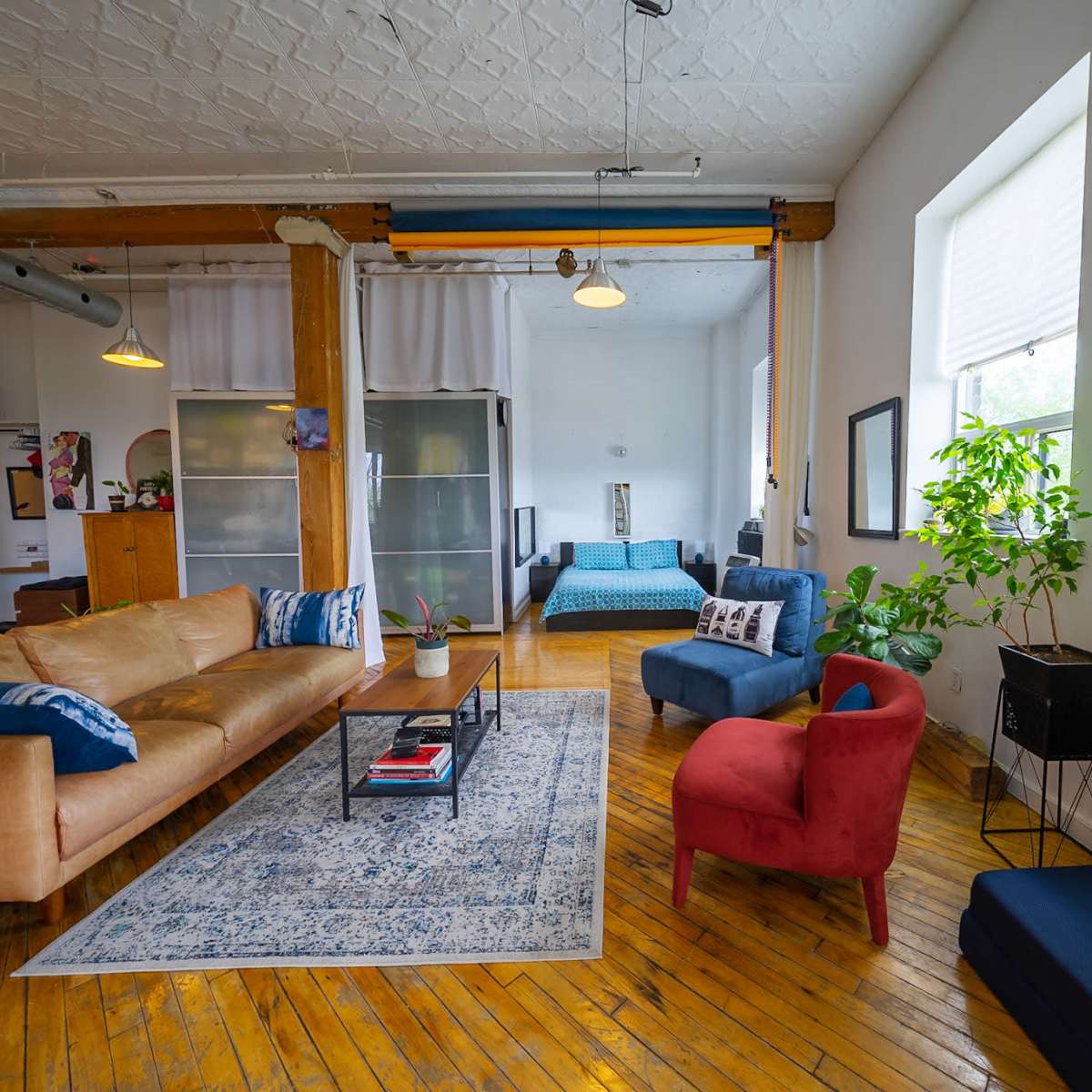 A spacious, light-filled apartment with wooden floors, featuring a large brown sofa, a blue chair, and a red chair, along with a coffee table and various plants.
