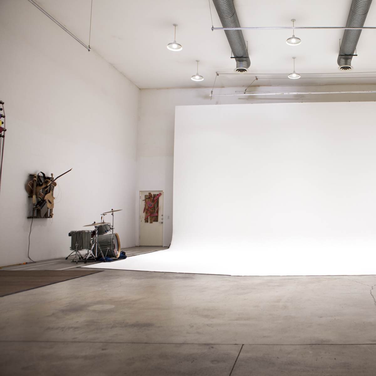 A large, empty studio space with white walls and a seamless white backdrop, featuring industrial lighting and a drum set in the corner.