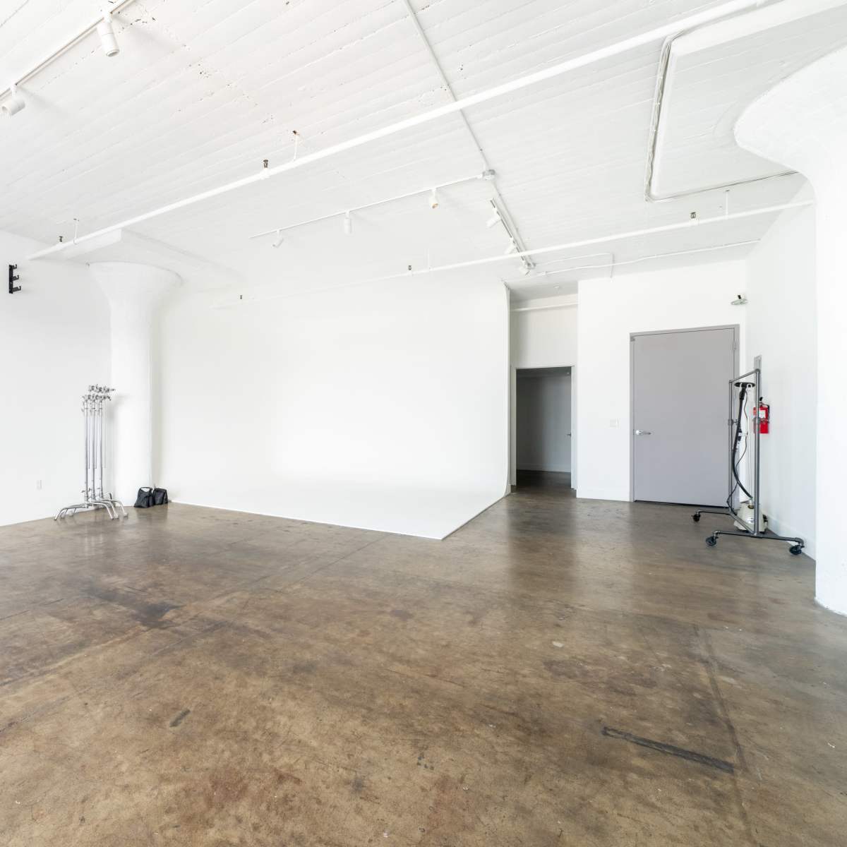 A spacious, empty studio with a white ceiling and walls, featuring concrete flooring and minimal furnishings.