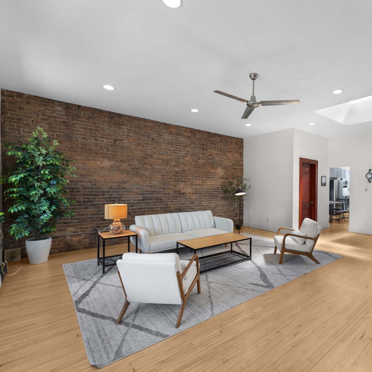 The room features a modern living area with a brick accent wall, a light-colored sofa, two chairs, a coffee table, a ceiling fan, and a large flat-screen TV mounted on a stand.