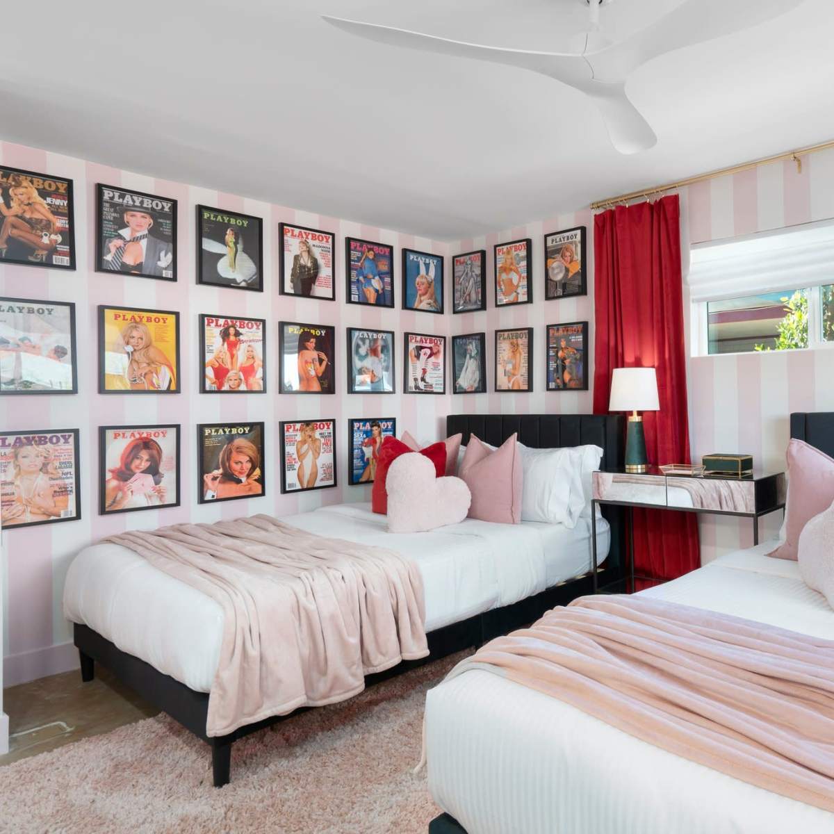A room with two beds, each adorned with pillows, surrounded by framed magazine covers on the walls and red curtains at the window.
