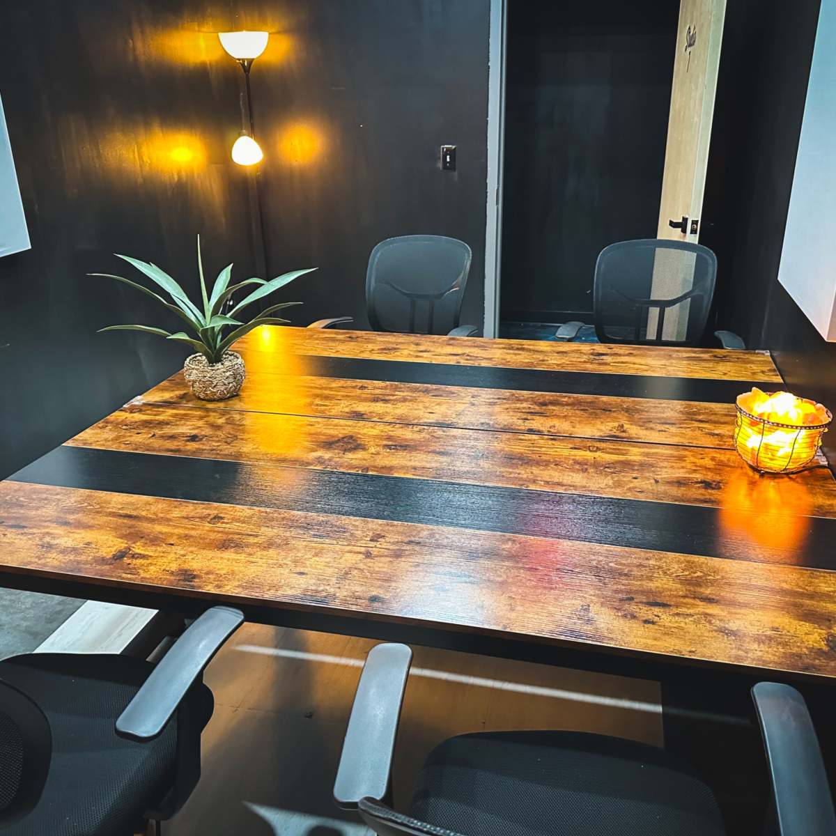 A wooden table with black stripes is set in a room featuring black walls, surrounded by four office chairs and decorated with a potted plant and a candle lantern.