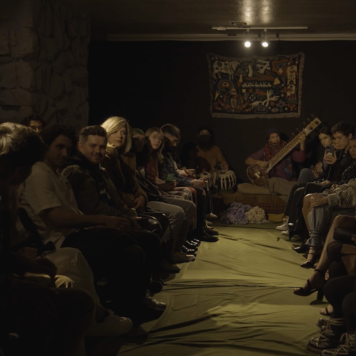 A group of people sits in a dimly lit room facing a musician playing a sitar, with a tapestry hanging on the wall behind them.