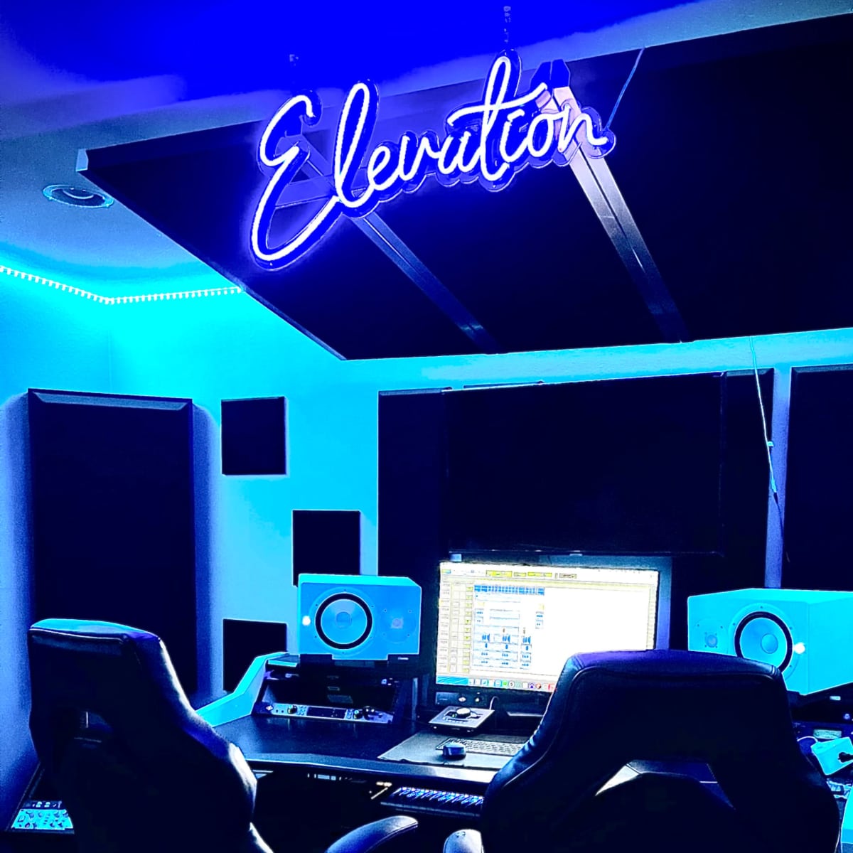 A music studio with two black chairs facing a computer setup, illuminated by blue lighting and a neon sign that reads "Elevation."