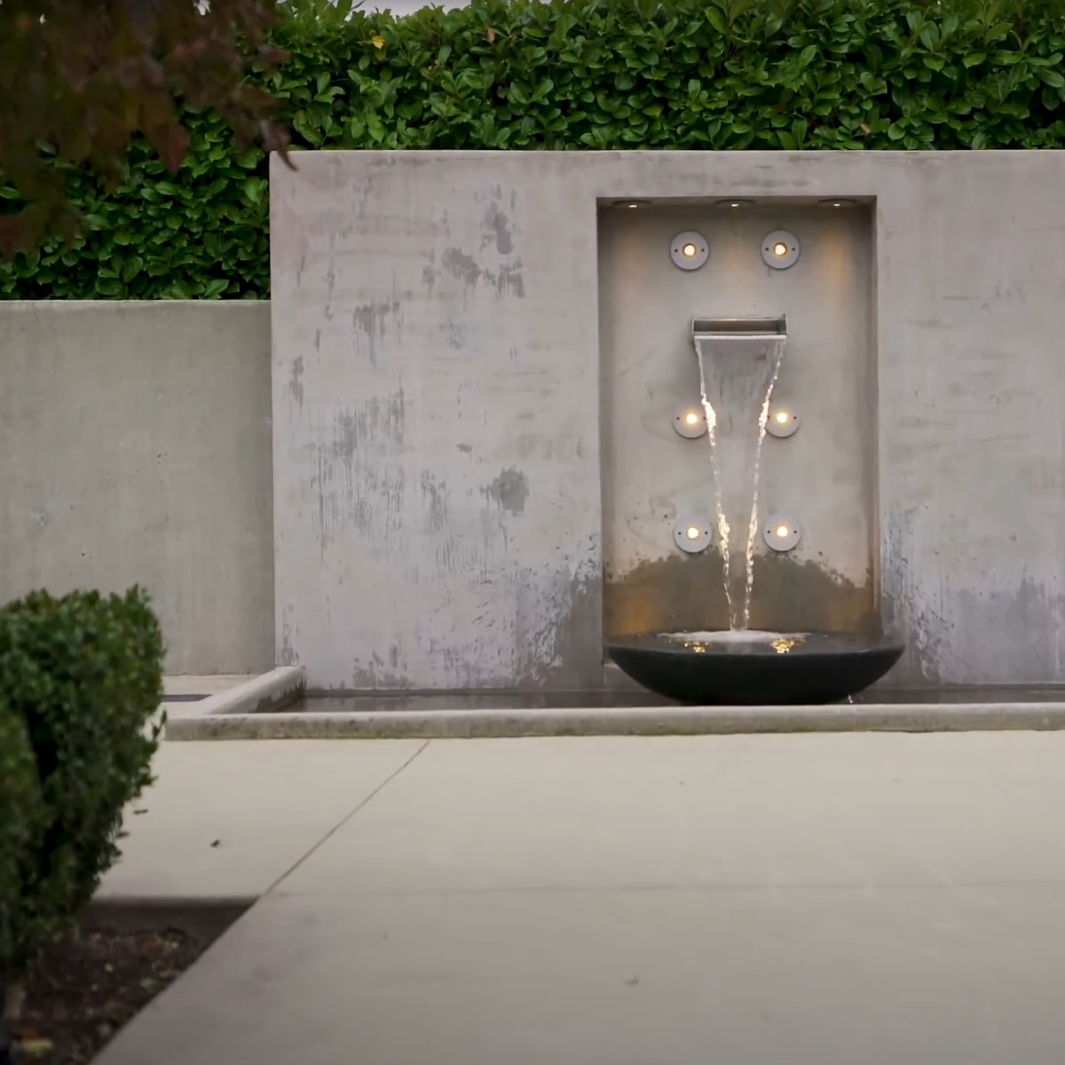 A modern water feature with a vertical wall, a fountain, and circular lights is situated in a landscaped area.