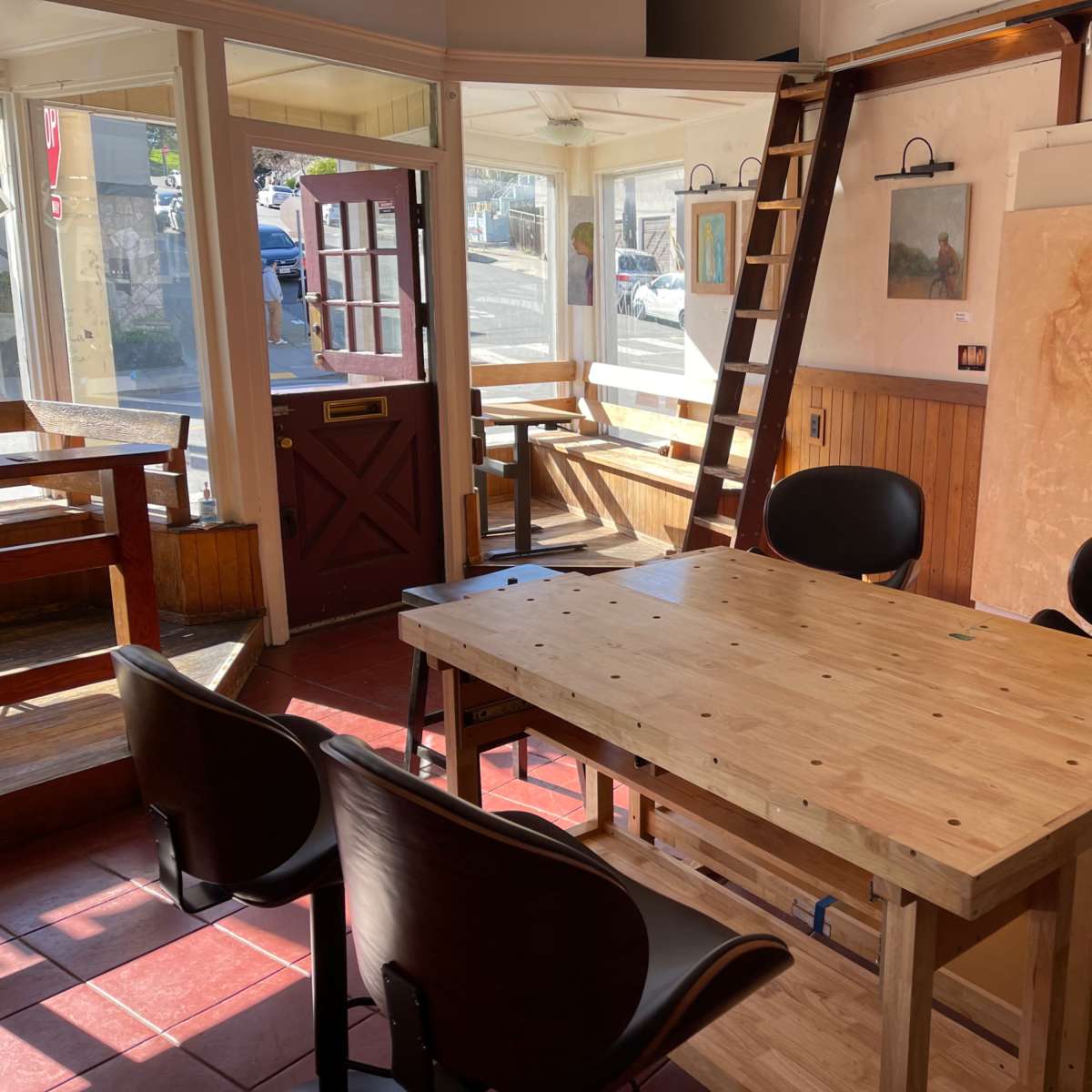 A sunlit art studio with a wooden table and chairs, featuring a ladder and paintings on the walls, along with a view of the street through a door.