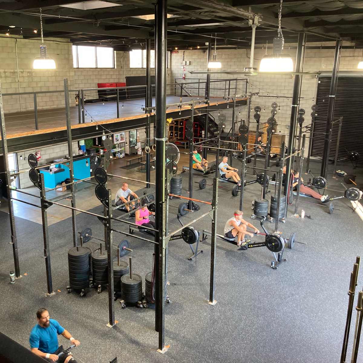 An indoor gym with multiple workout stations, including weights and rowing machines, occupied by several individuals exercising.