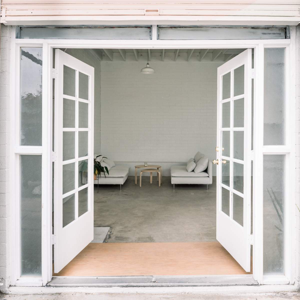 A pair of white double doors opens to a spacious, minimal interior with two sofas and a small table, flanked by potted plants.
