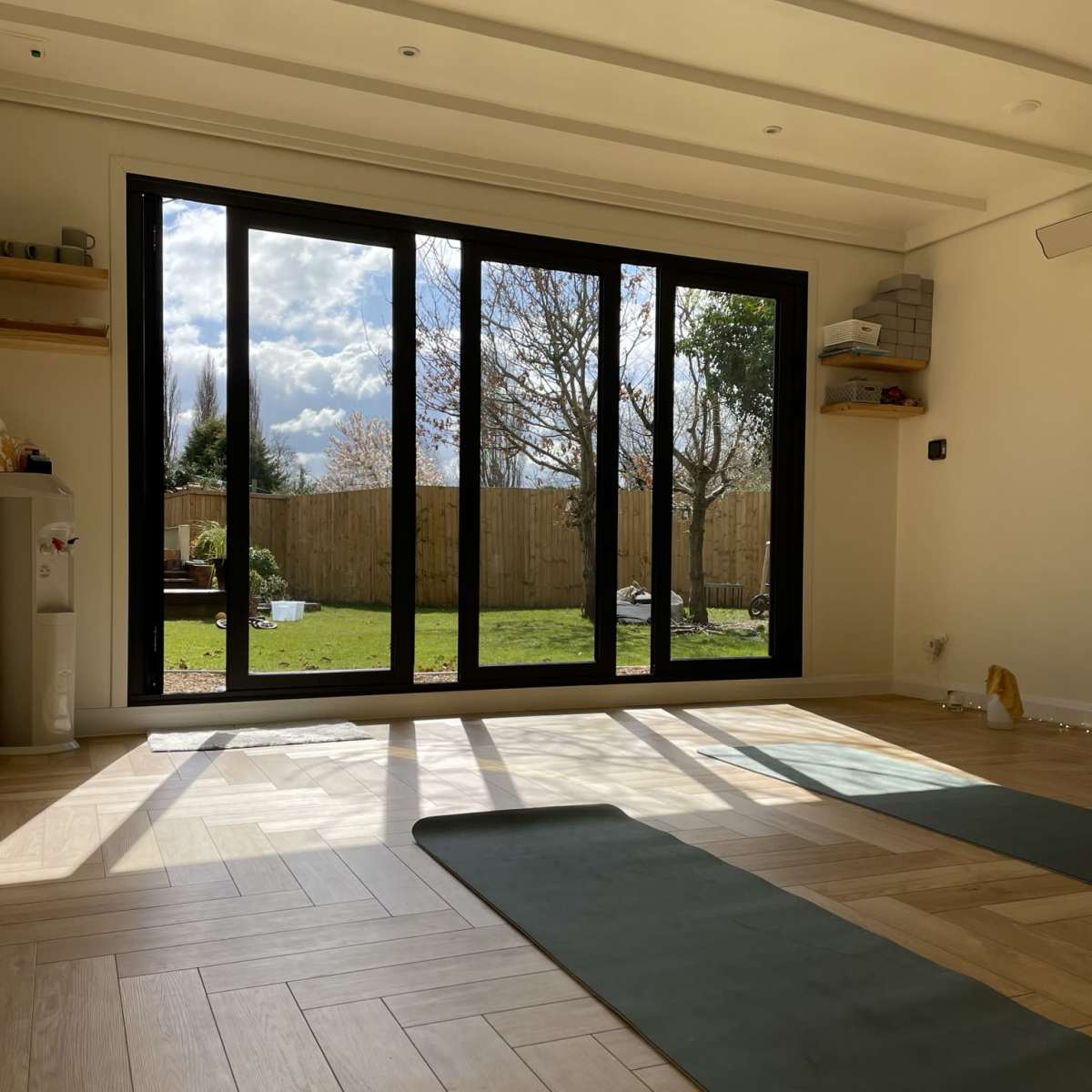 A bright room with large black-framed windows overlooks a green yard with trees and a partially cloudy sky.