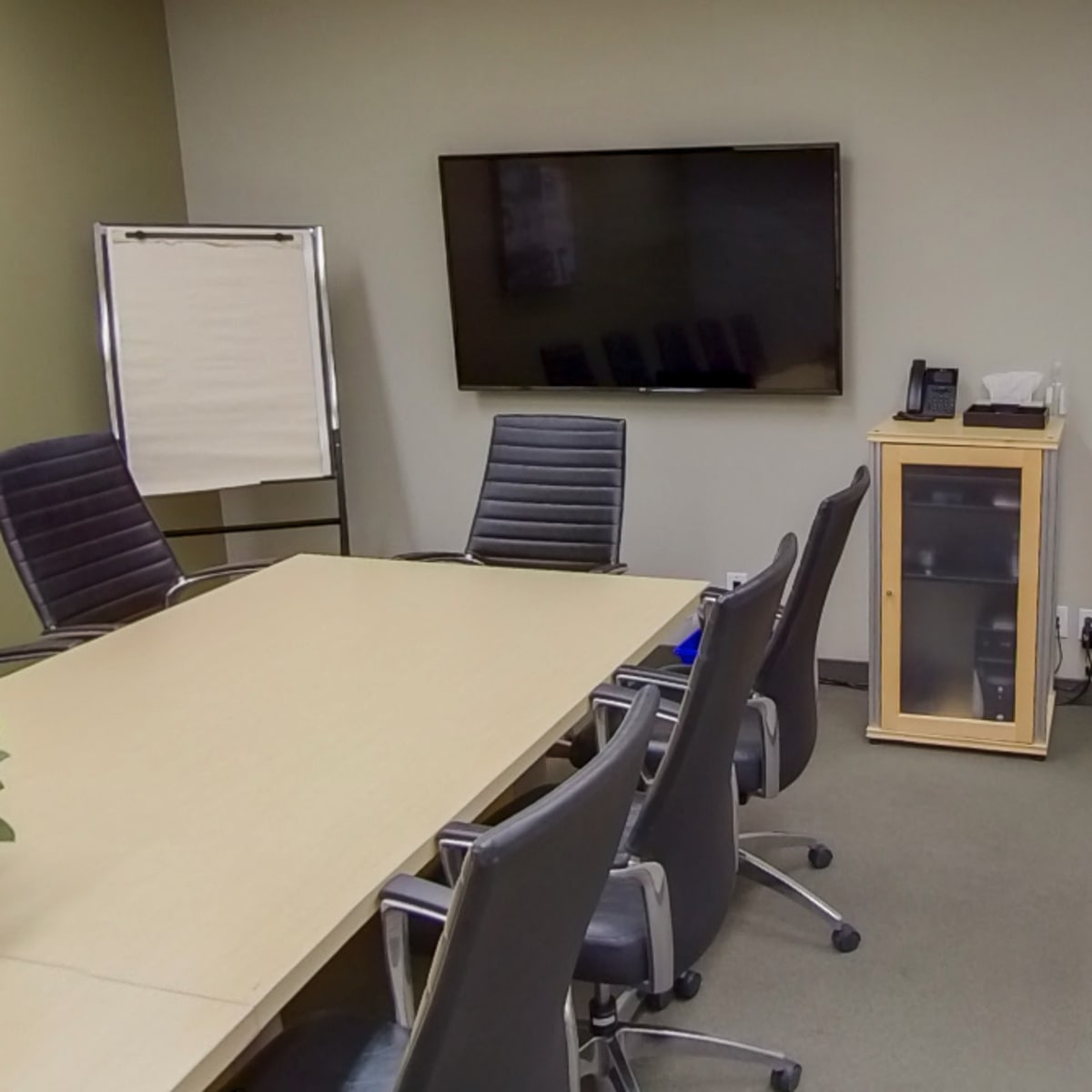 A conference room features a long table, several rolling chairs, a large television mounted on the wall, and a small cabinet in the corner.