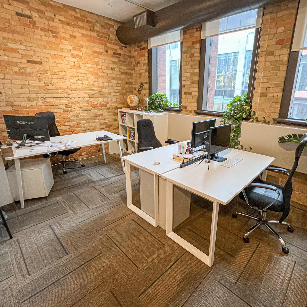 A modern office space with two desks, plants, large windows, and exposed brick walls.