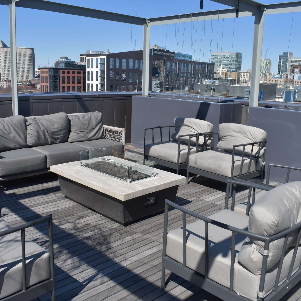 A rooftop lounge area with gray seating arrangements surrounding a central fire table, overlooking a city skyline.