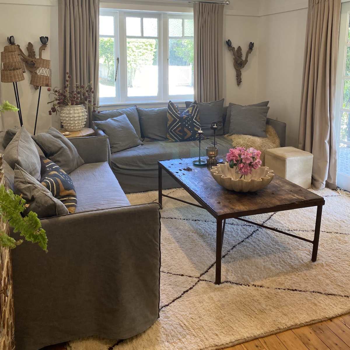 A modern living room featuring a large gray sectional sofa, a wooden coffee table with a bowl of flowers, and large windows dressed with light curtains, with a plush rug on the wooden floor.