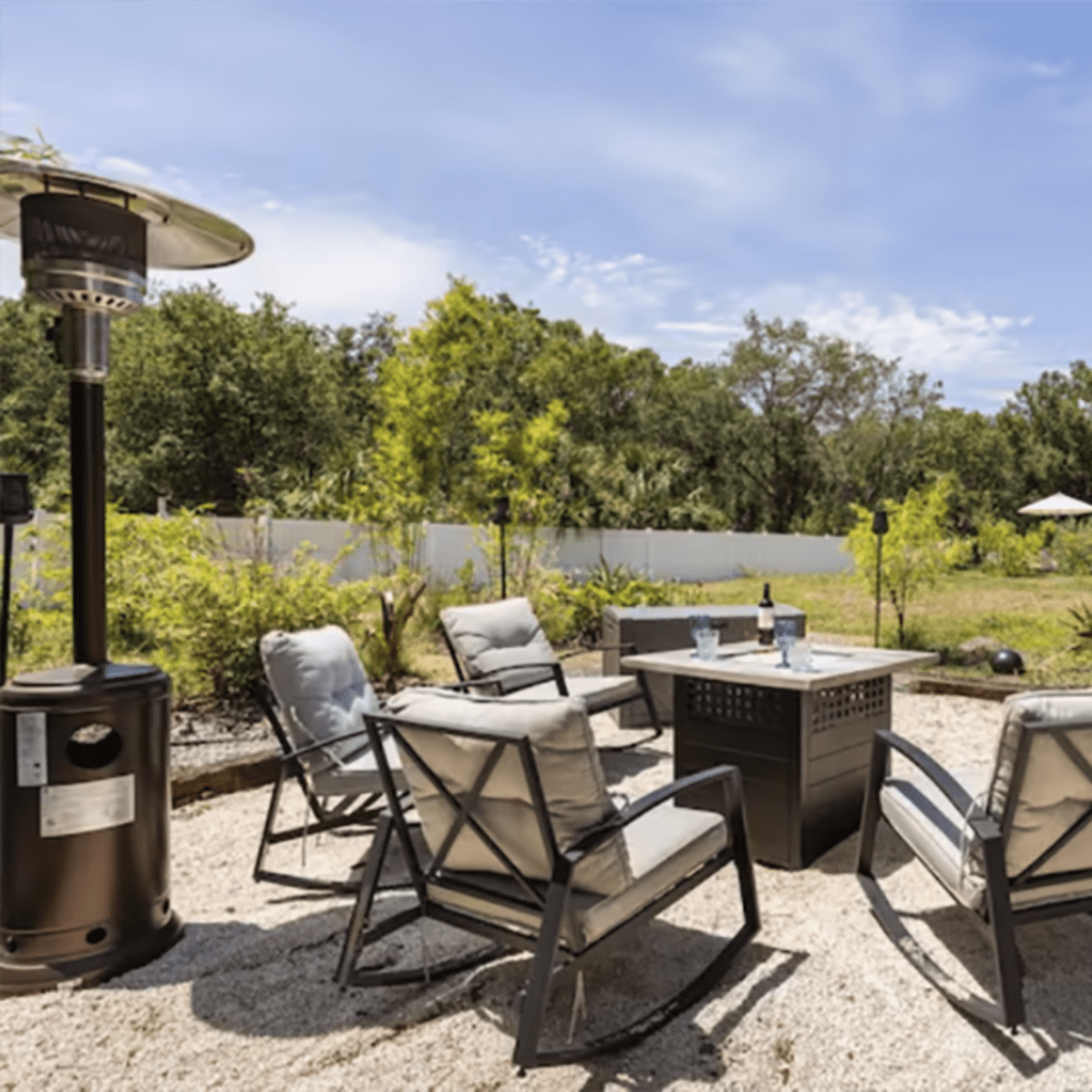 A gravel patio features four chairs arranged around a central table with a portable heater nearby, surrounded by greenery and outdoor furniture in the background.