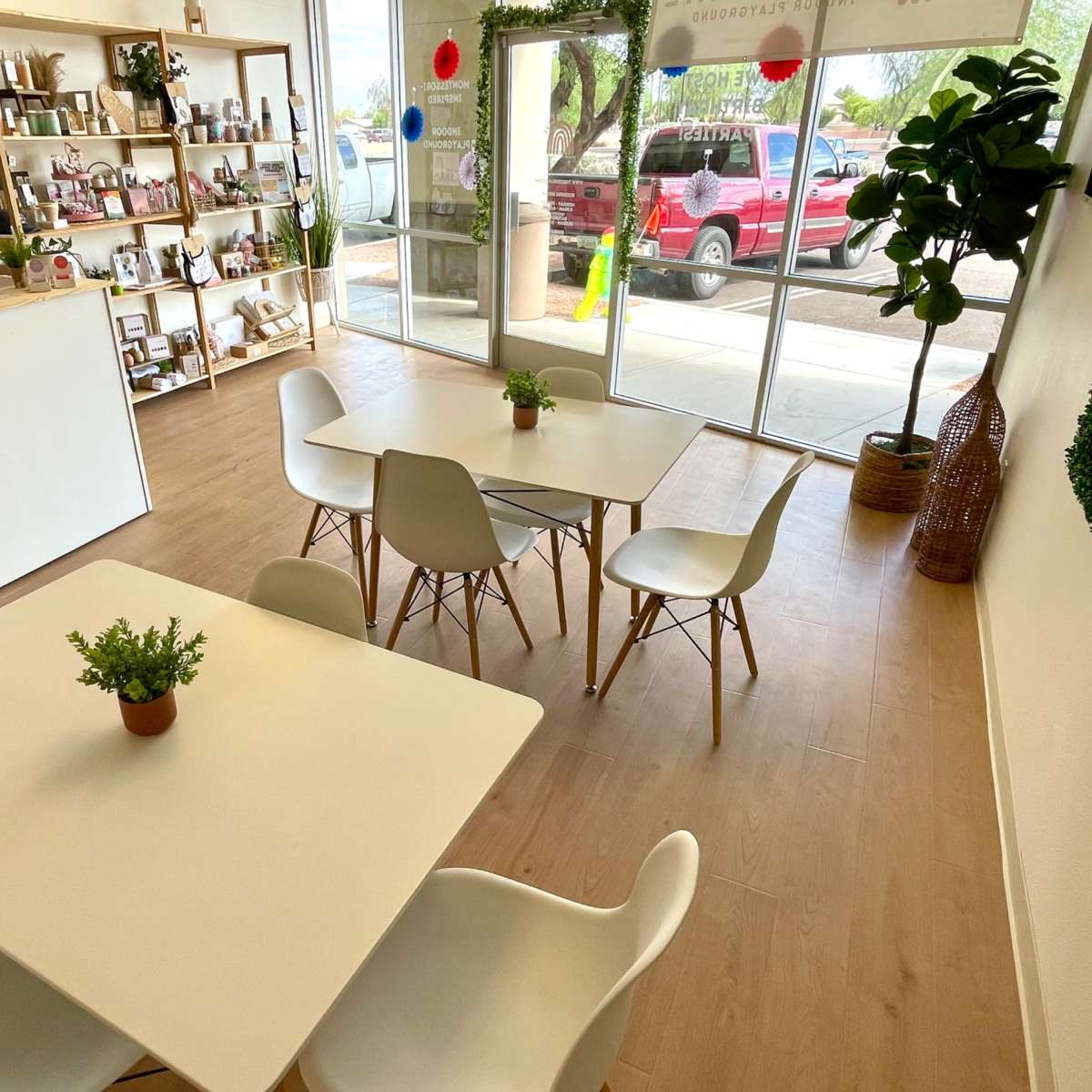 A bright interior of a café featuring wooden flooring, white tables surrounded by chairs, and decorative shelves with various items in the background.