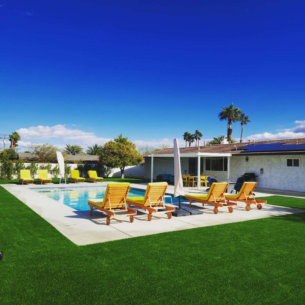 A rectangular swimming pool is surrounded by yellow lounge chairs and artificial grass under a clear blue sky.