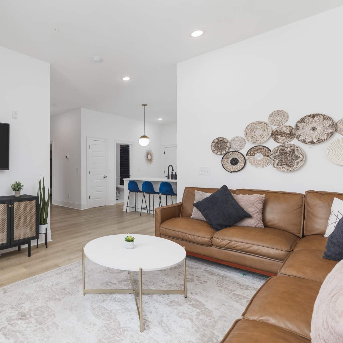 A modern living room features a brown leather sofa, a round coffee table, a television on a console unit, and decorative woven wall art.