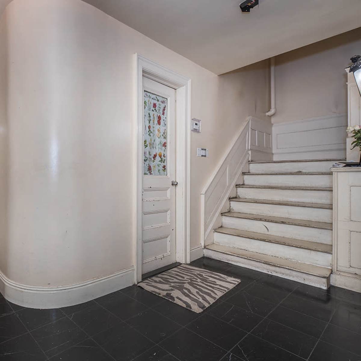 A hallway with a staircase on the right, a door near the bottom of the stairs, and a patterned runner on the black tiled floor.