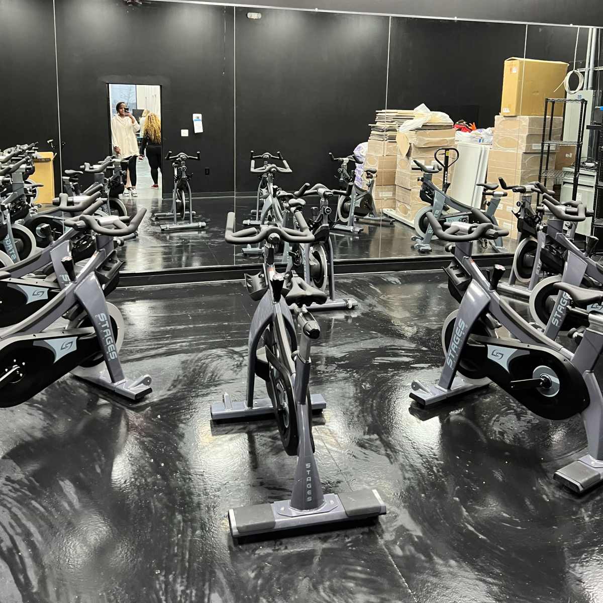 A spacious indoor cycling room with several stationary bikes and a large mirror reflecting the entrance and boxes stacked in the corner.