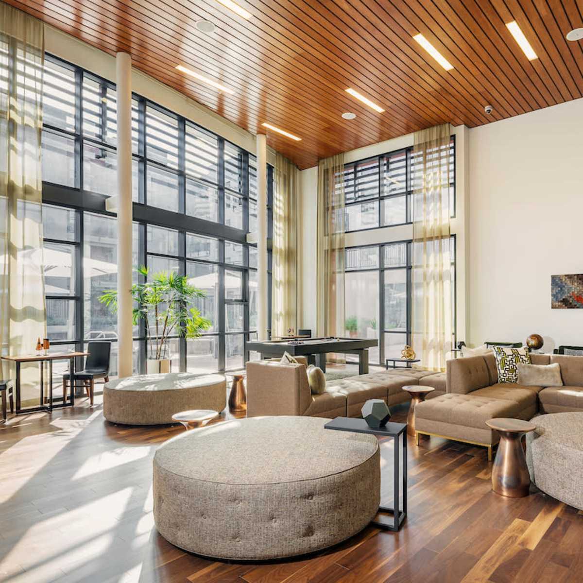 A spacious modern living area features large windows, light wood ceilings, and a combination of round and angular furniture with a neutral color palette.
