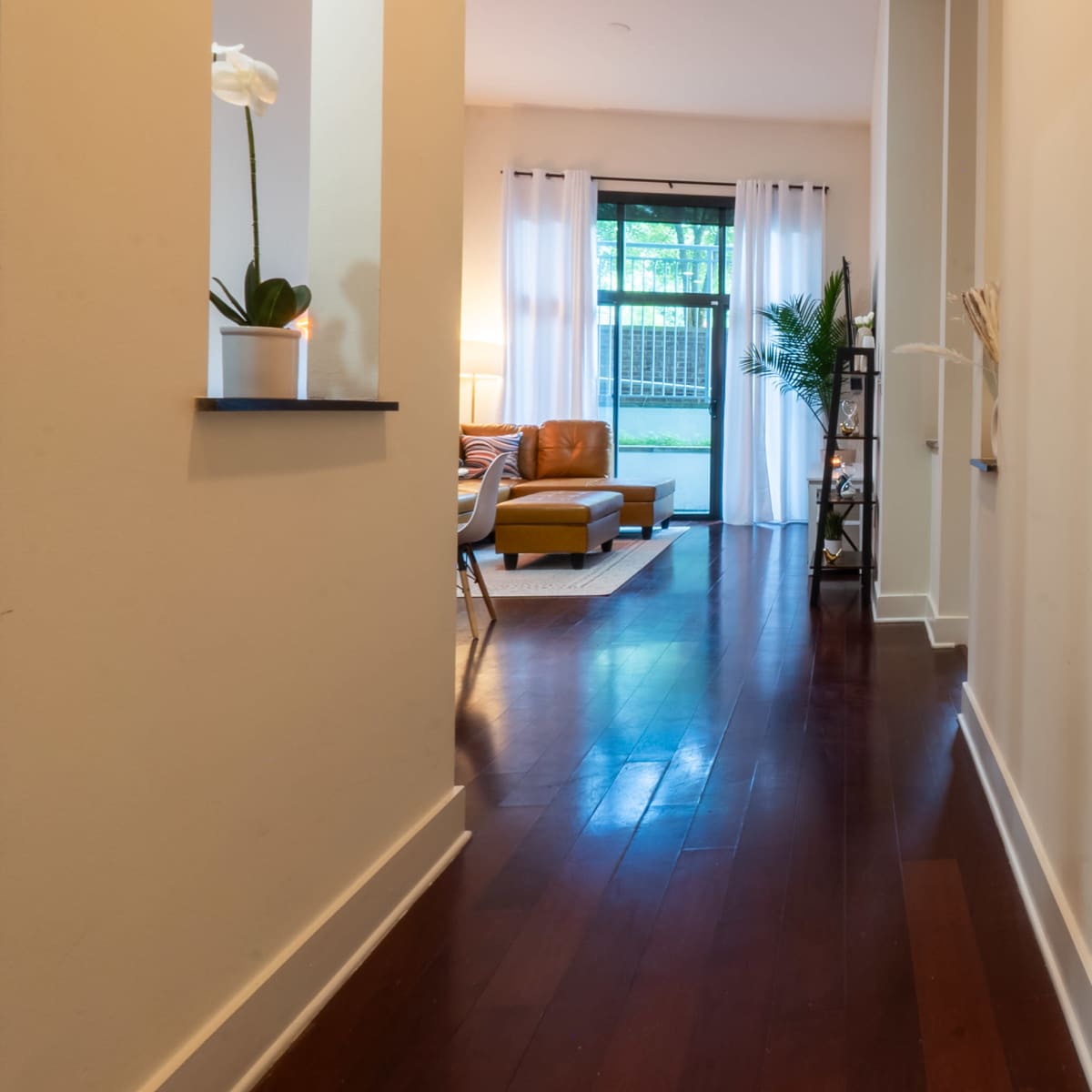 A hallway with hardwood floors leads toward a cozy living room with large windows and light-colored curtains.
