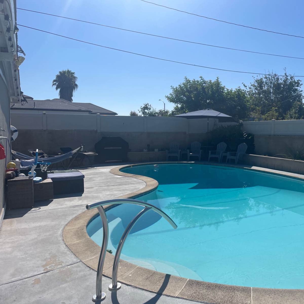 A clear blue swimming pool surrounded by a concrete deck and several chairs, with a palm tree in the background.