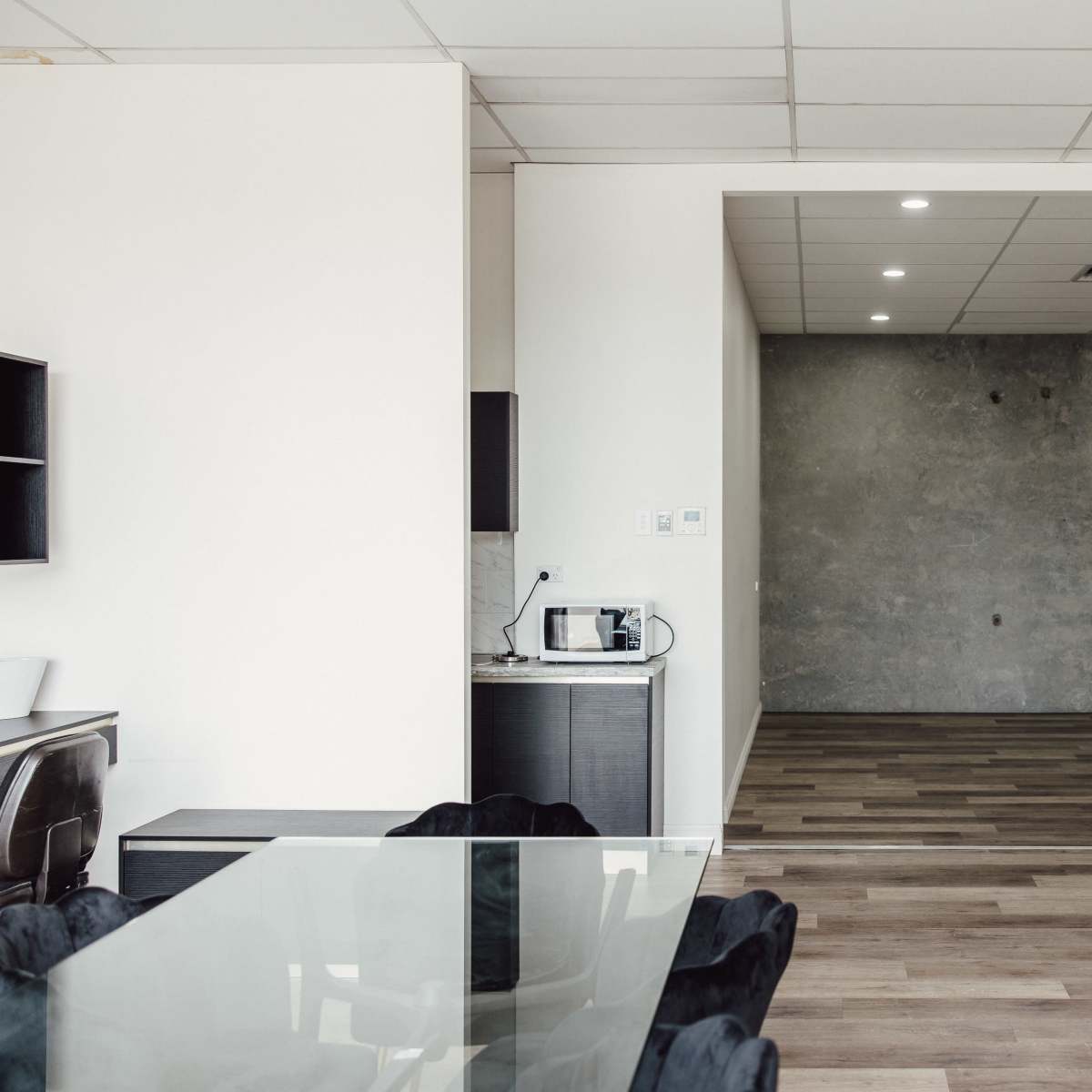 A modern office space features a dining area with a glass table and black chairs, leading to a minimalistic room with a concrete wall and wooden flooring.