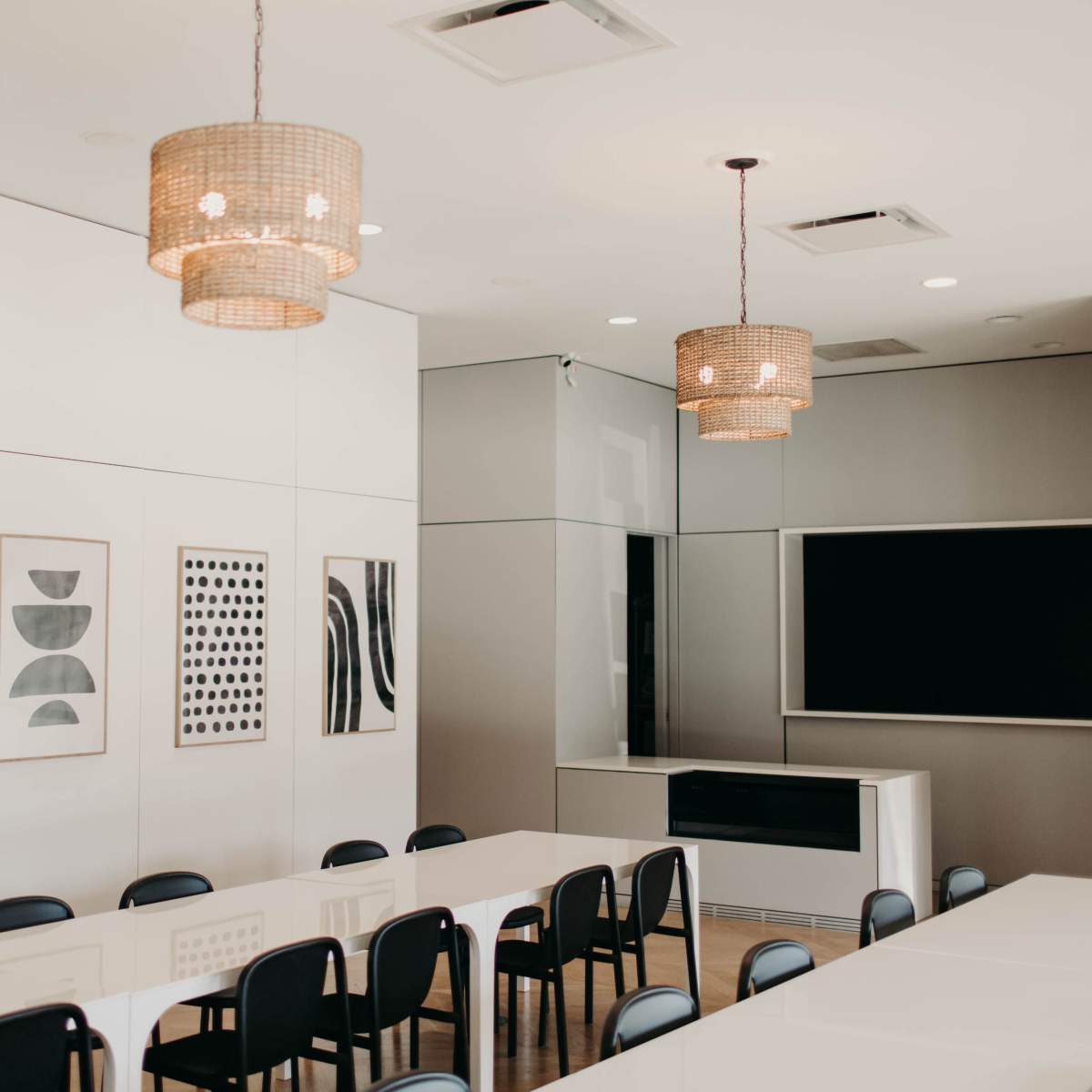 A modern conference room featuring a long white table, black chairs, and framed artwork on the walls, illuminated by woven pendant lights.
