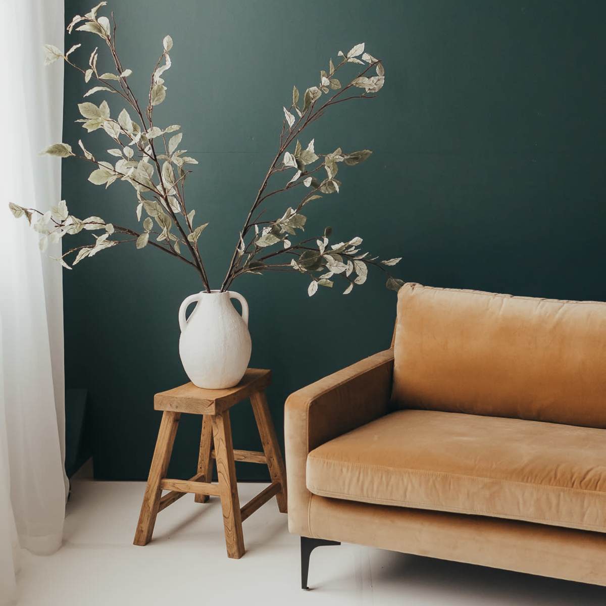A brown velvet sofa is positioned next to a wooden stool holding a large white vase with branches against a dark green wall.