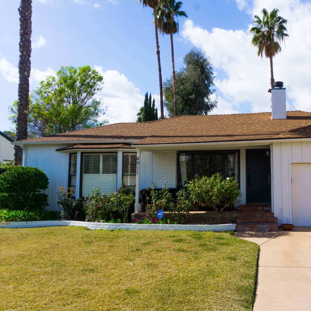 A single-story suburban house with a brown roof and a driveway leading to an attached garage, surrounded by green landscaping and palm trees.