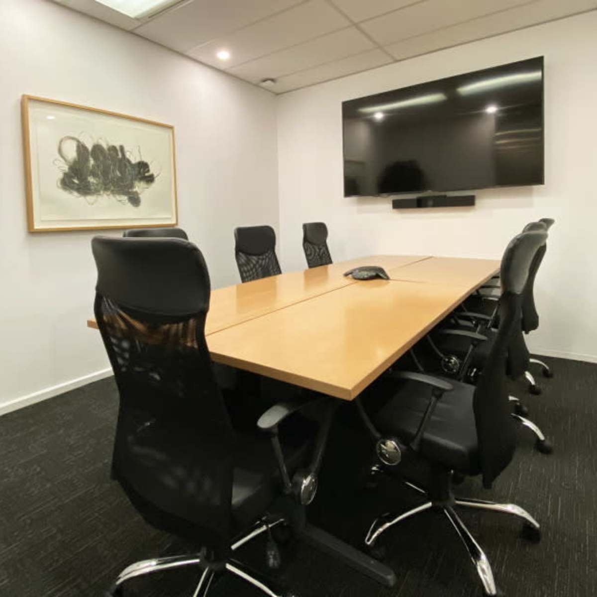 A modern conference room with a long wooden table surrounded by black ergonomic chairs and a large flat-screen monitor on the wall.