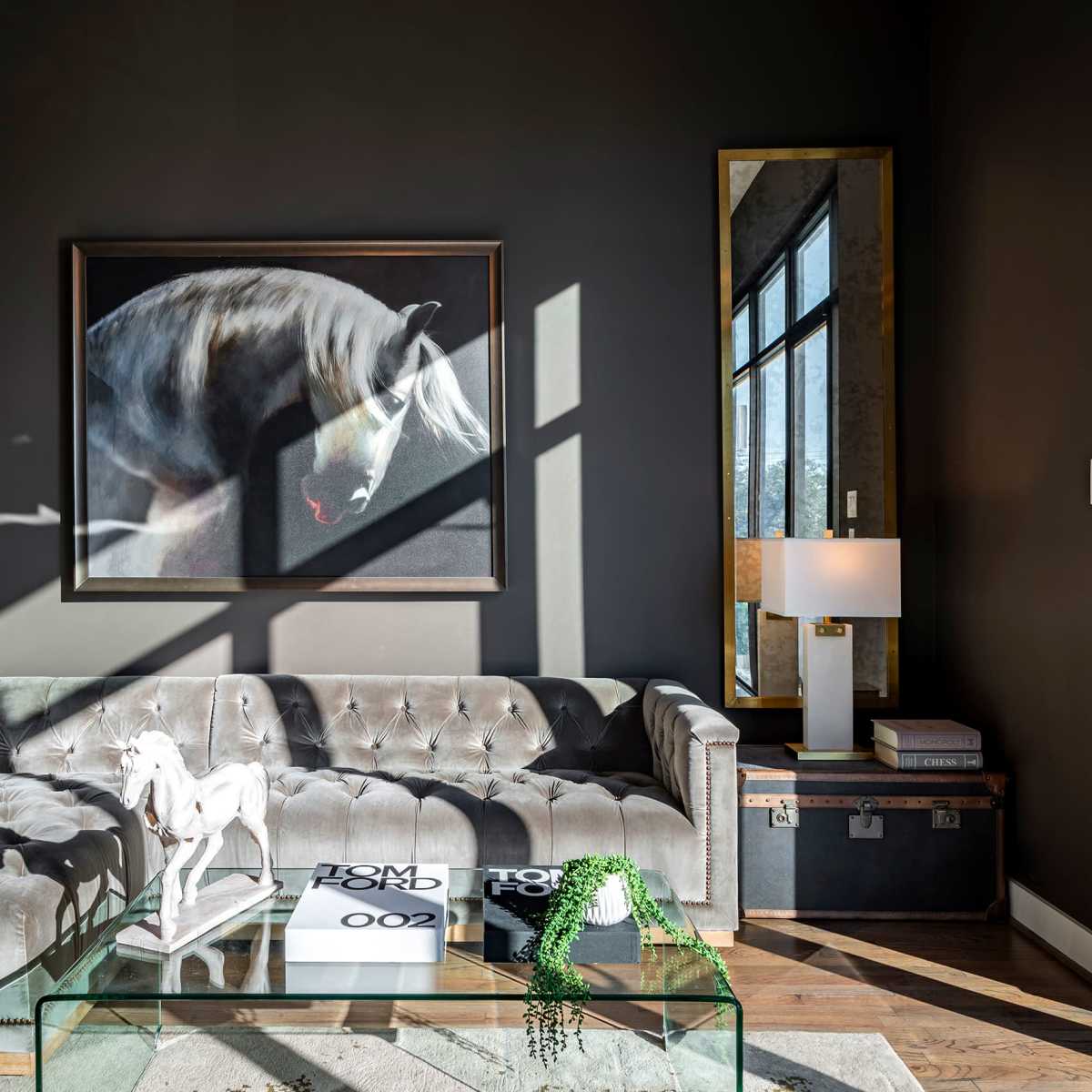 A modern living room features a tufted gray couch, a glass coffee table, and a large painting of a horse hanging on the wall.