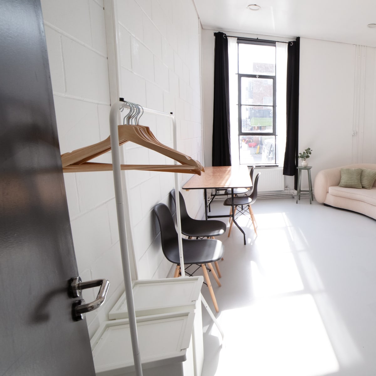 The room features a sofa, a table with chairs, a small cabinet, and several plants, all arranged under large windows with natural light.