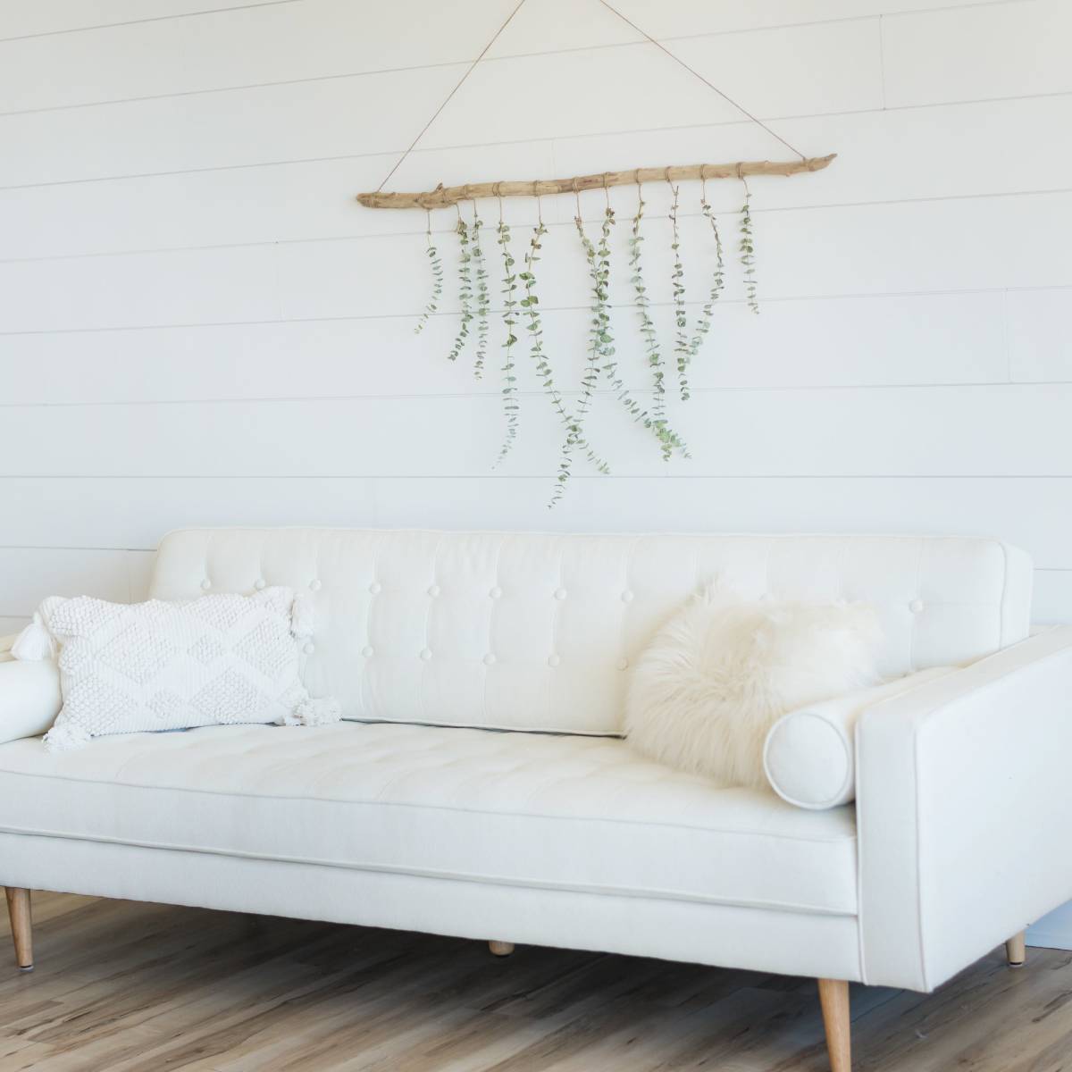 A white sofa with decorative cushions sits against a light wooden wall, accompanied by a round wicker side table and a hanging plant display.