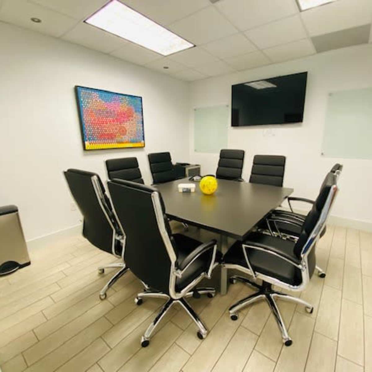 A conference room features a large table surrounded by black chairs, a television on the wall, and a piece of art above the table.