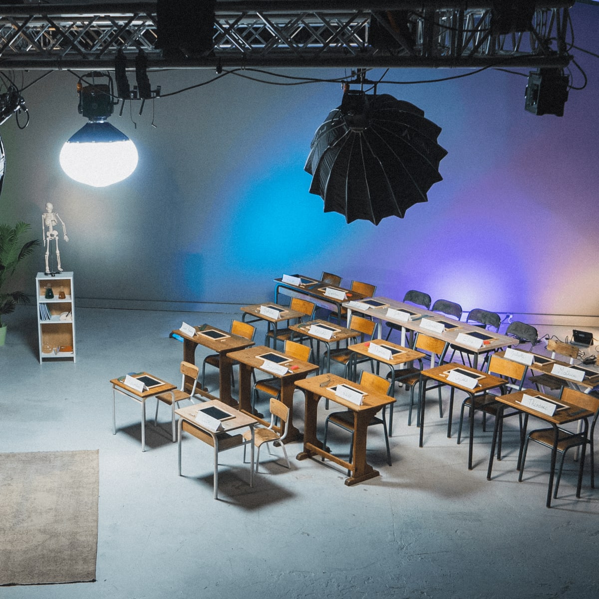 A classroom setting features rows of wooden desks and chairs, a blackboard, a globe, and studio lighting hanging from the ceiling.