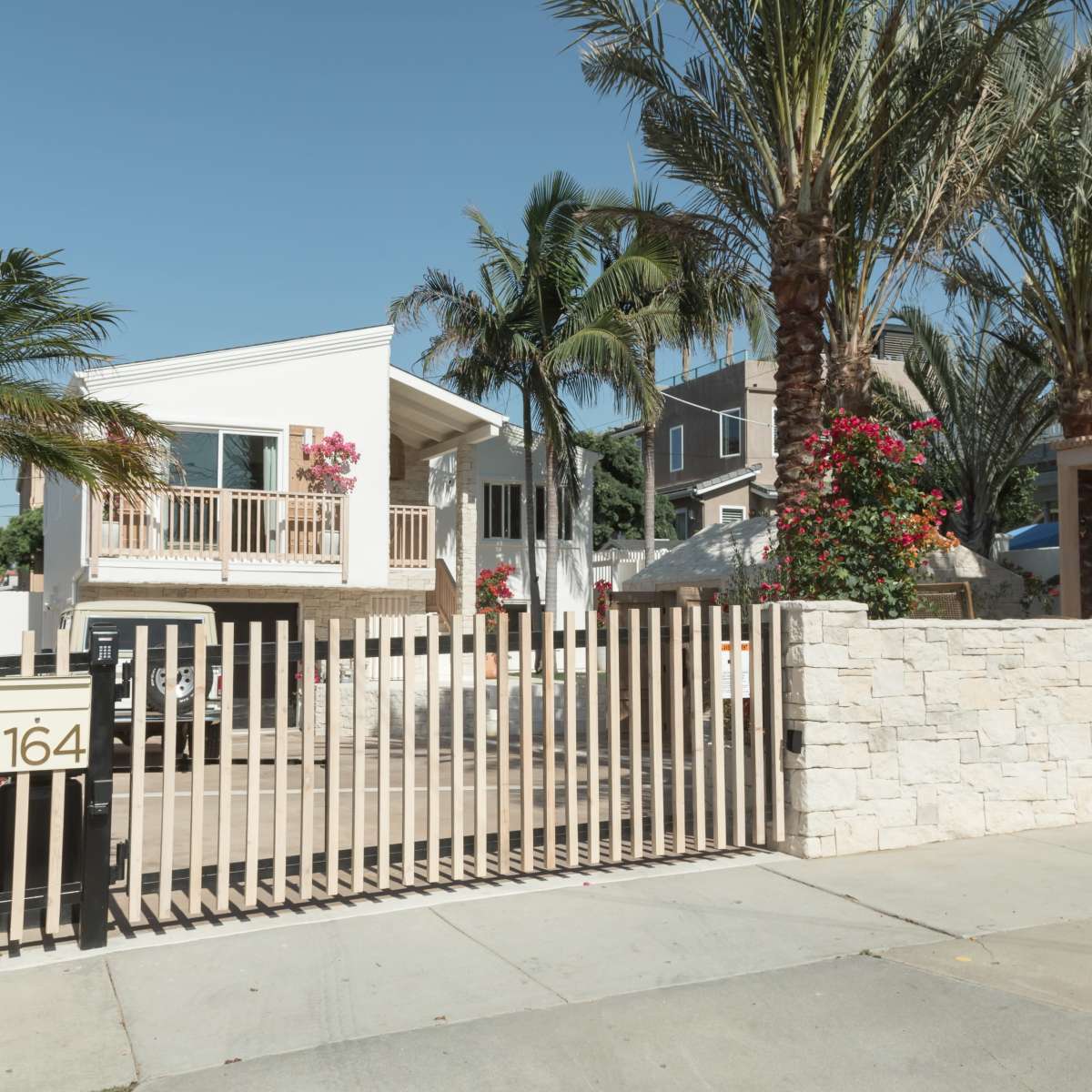 A modern house with a balcony and flowering plants is enclosed by a stone wall and a wooden gate.