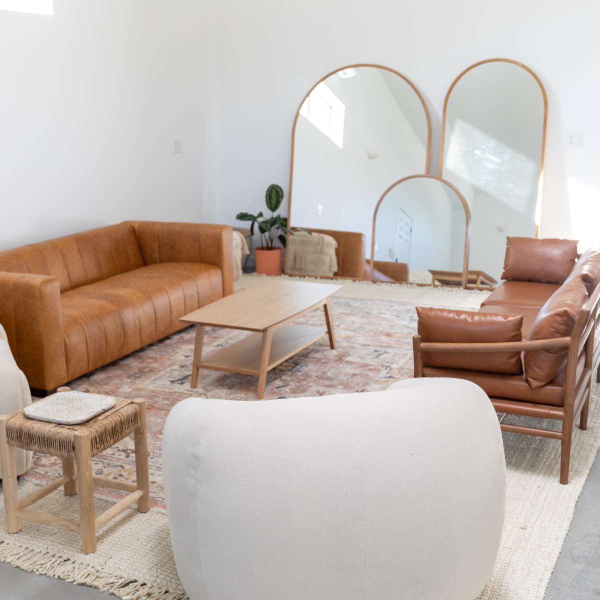 A modern living room features a brown leather sofa, light-colored armchairs, a wooden coffee table, and a patterned area rug, all arranged around large mirrors on a white wall.