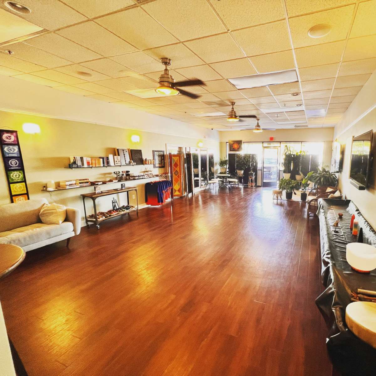 A spacious, well-lit room with wooden floors, featuring soft seating, various instruments, and shelves lined with items for a wellness or meditation environment.