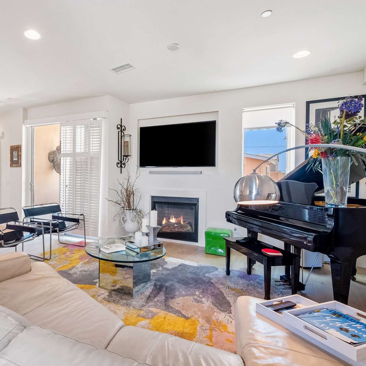 A modern living room features a white leather sofa, a grand piano, a glass coffee table, and a fireplace, with light streaming in through large windows.