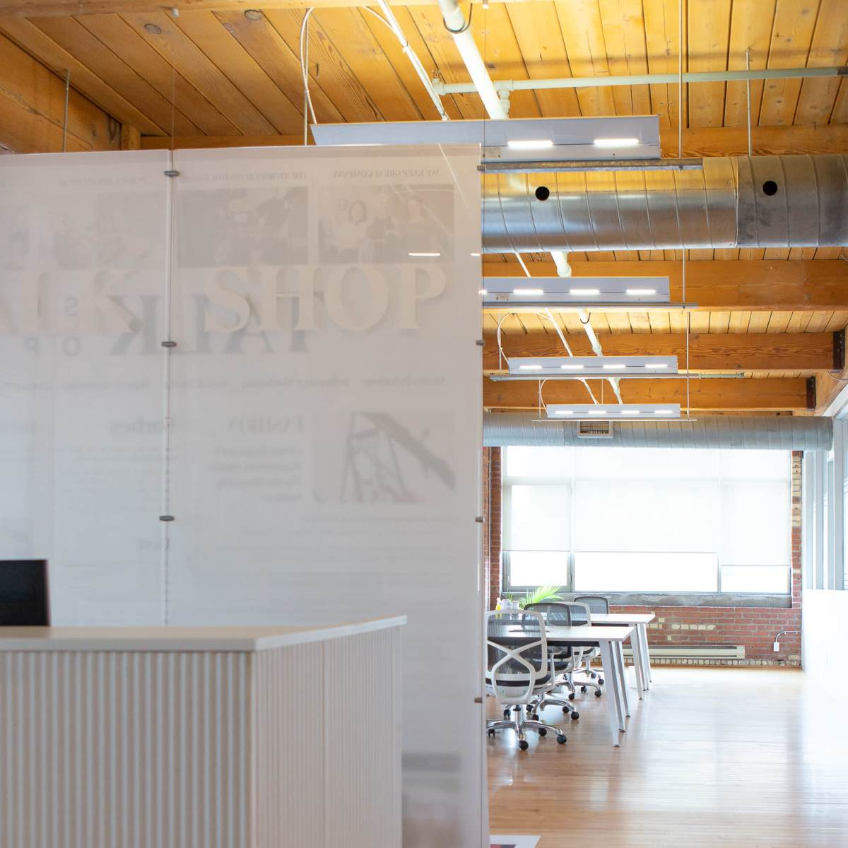 A modern office space with wooden floors, large windows, and a partition separating the reception area from the workstations.