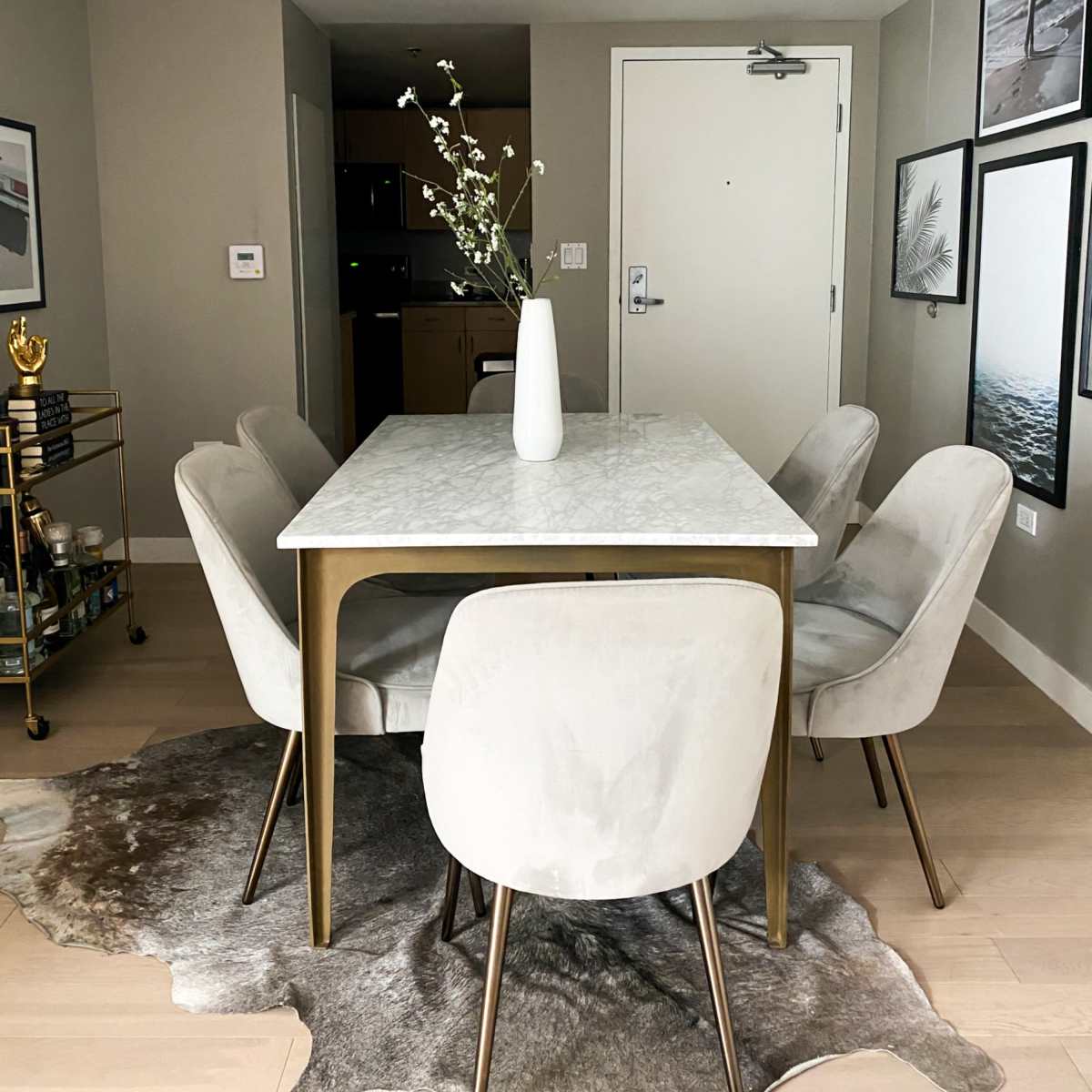 A dining area features a marble-topped table surrounded by six light-colored chairs, with a vase of flowers as the centerpiece and framed images on the wall.