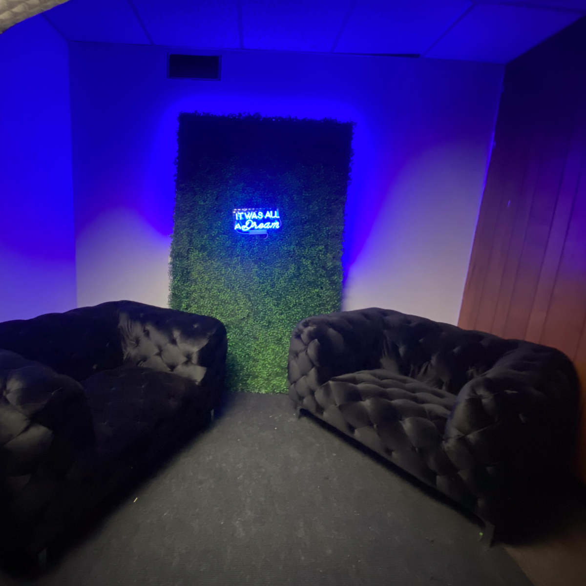 A small lounge area with two black tufted sofas facing a green wall with a neon sign that reads "It was all a dream."