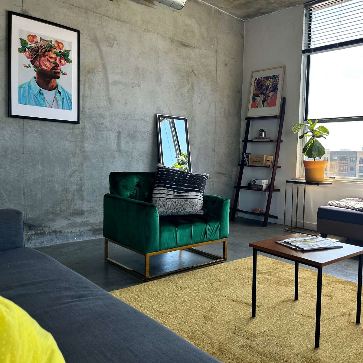 A modern living room featuring a green velvet chair, a wooden coffee table, a large window with natural light, and a mix of artwork on the walls.