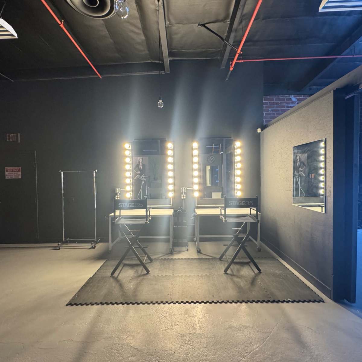 A dimly lit room with two illuminated makeup mirrors, each accompanied by a director's chair, and a large window providing natural light.