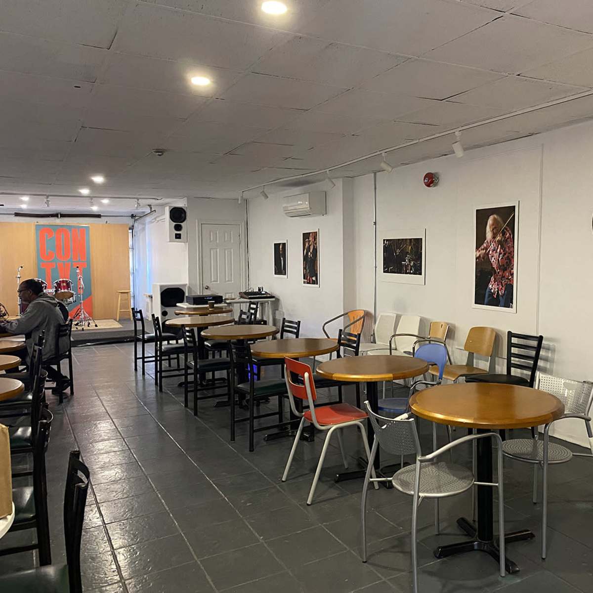 A spacious café with several tables and chairs arranged around the room, featuring wall art and a small stage at one end.