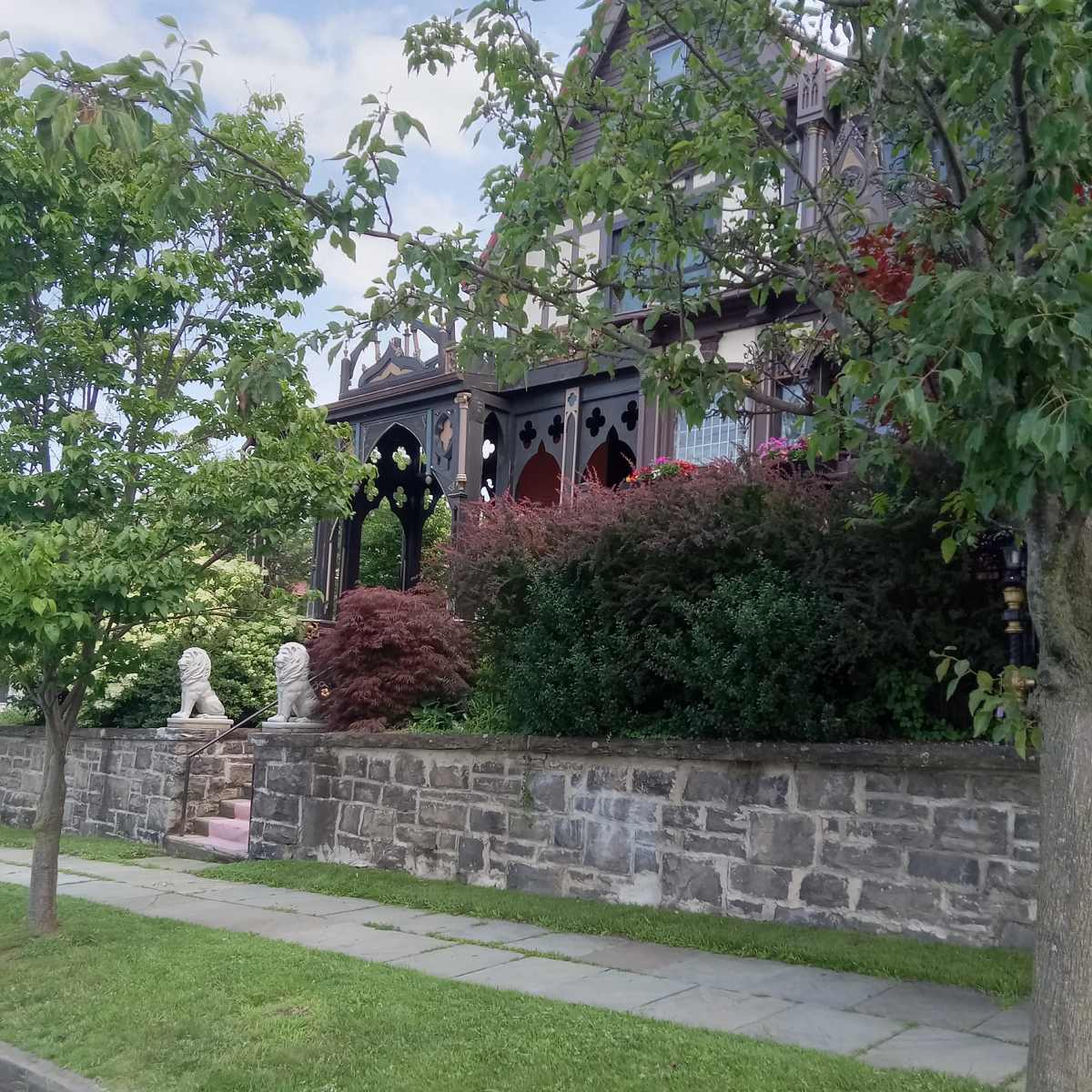 A large, Victorian-style house with a elaborate porch, surrounded by trees and a stone wall.