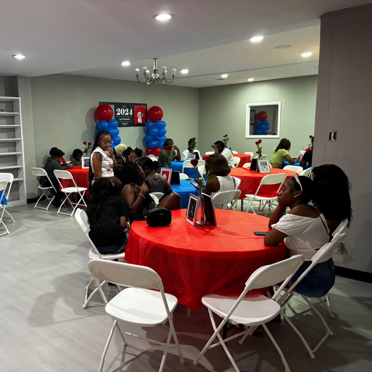 A group of people sits at red and blue table settings in a decorated room, with a banner in the background indicating the year 2024.